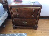 Front view of wood side table showing three drawers with ornate brass handles and brass trim details, on hardwood floor with adjacent bed.
