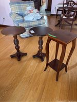 Three wood tables: two round pedestal tables and one square side table with lower shelf. Placed on wood floor, close to patterned armchair.