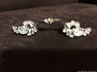 Front view of decorative dark cream and gold embossed metal drawer pull showing detailed floral and scroll motifs.