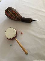 Photo showing the gourd shaker painted with natural and red colors alongside the small hand drum with a white drumhead, wooden handle, and noise makers attached by beads on strings.