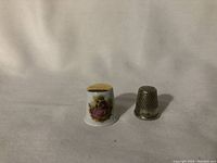 Two thimbles side by side on a beige cloth; porcelain thimble decorated with pink flowers and gold top, metal thimble plain silver color with holes.