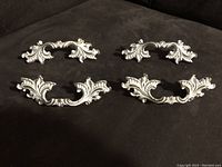 Four decorative drawer pulls arranged on fabric surface showing front view with cream color and gold embossed details