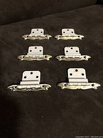 Top view of all six cream colored metal and gold embossed drawer pulls arranged on dark fabric showing design and hinge.