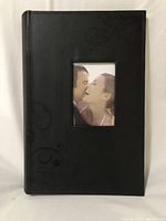 Front cover photo of Uniek photo album showing black faux leather with swirl etching and 3 photo openings with a couple's photograph inside the center window.