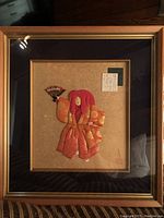 Front of framed artwork showing padded figure wearing bright orange traditional Japanese attire, holding a fan, mounted on beige textured background with decorative mats and wooden frame.