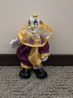 Front view of clown doll with porcelain head, dressed in purple and yellow satin outfit with ruffled collar.