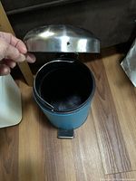 Front angled view showing the open silver metal lid, teal body, black removable inner container with wire handle and foot pedal.