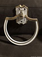 Front view of the cream colored metal towel holder with gold embossed accents showing the semi-circular ring and mounting plate