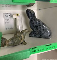 Three stone animal figurines on shelf including eagle head, dark seal, and green stone bird with a size note taped nearby.