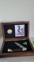 Open wooden box showing the folded cutter knife in the bottom compartment, and the bear coin alongside a duck illustration in the top compartment.