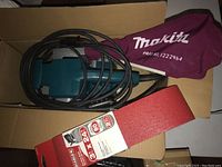 Open carton showing teal Makita belt sander with power cord, maroon Makita dust bag, and spare sanding belts package