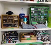 Two white shelves containing a wooden drawer organizer with sewing supplies, a green plastic organizer with several small compartments filled with sewing notions, and multiple spools of thread on racks.