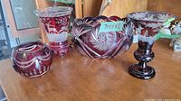 Four cranberry and clear glass pieces displayed on wooden surface: two detailed vases, one large bowl, one small bowl.
