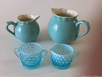 Photo showing the two blue ceramic milk jugs and the small light blue hobnail glass creamer and sugar set.