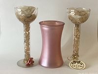 Pair of clear glass candlesticks with gold and fabric rose adornments flanking a matte pink La Crème vase