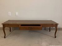 Long solid wood coffee table with a single shallow drawer open, showing interior and metal pull handle. Medium brown finish and cabriole legs visible.