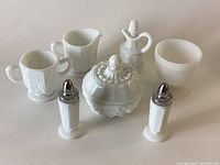 All six milk glass pieces arranged together showing the covered dish, salt and pepper shakers, creamer, sugar bowl, and flower pot under natural light.