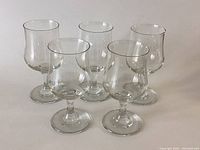 Five clear stemmed glasses arranged in two rows on a white surface, showing rounded bowls and short stems.