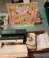 Photo showing a box with various books including 'Tender Is the Night' by F. Scott Fitzgerald, a green hardcover book, a stack of small biography booklets, the 1995 Beatrix Potter Peter Rabbit calendar, and other books.