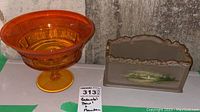 Orange glass pedestal bowl with flared rim next to ornate glass planter with painted lake scene and gold trim.