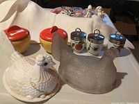 Full view of all items including two ceramic lidded jars, three porcelain egg coddlers with stainless steel lids, milk glass hen on nest and clear glass hen on nest lids.
