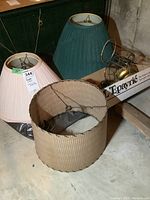Photo showing two pleated fabric lamp shades in pink and green, the large beige cylindrical pleated lamp shade, and box of assorted metal lamp parts.