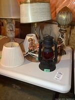 Photo of the lot showing Dietz oil lantern, ceramic lamp with Hummel-style figures, metal cherub lamp with shades on a white table.