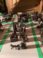 Overview of metal miniatures arranged on table with green tape grid, showing a variety of designs including dinosaurs, engines, and architectural models.