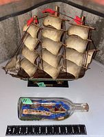 Photo shows multi-mast whaling ship model with cream sails and red flags alongside a glass bottle with three ships inside set on a blue base. A ruler for scale is visible at the bottom.