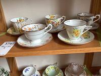 Five sets of vintage teacups and saucers arranged on wooden shelves, showing floral designs and gold trim