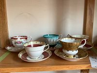 Five vintage teacup and saucer sets displayed on a wooden shelf, showing various design patterns and colors including red, turquoise, and gold accents.