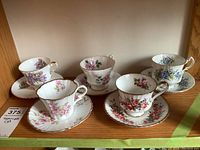 Five fine bone china teacup sets with saucers displayed on a wooden shelf, showing various floral patterns and gold trim