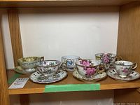 Six tea cup and saucer sets arranged on a wooden shelf showing various floral patterns and gold accents.