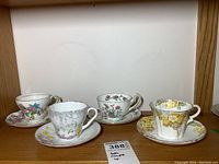 Four porcelain teacup and saucer sets displayed on a wooden shelf, showing distinct floral patterns from different makers.