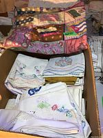 Box containing mostly white and off-white cotton pillow cases with colorful floral embroidery and a folded flannel patchwork blanket on top.