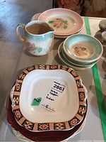 Photo showing the jug with floral design, cereal bowls stacked, pink rim dinner plates, and geometric border dinner plates underneath, all part of the set.