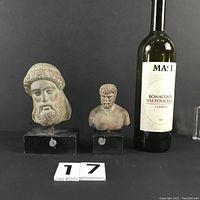Two replica Greek head busts on black wood bases displayed with wine bottle for scale