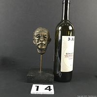 Frontal view of bronze head sculpture next to a wine bottle for scale, showing expressive facial details and stone base.