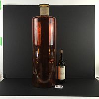Large amber-colored painted glass vase or bottle approximately 30 inches tall standing next to a wine bottle for scale. Neck wrapped with twine and metallic rim. Visible scratches on painted surface.