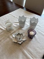 Top-down view of five assorted small candles and holders on a beige fabric surface, showing two white metal pierced holders, one white ceramic flower holder, one purple glass votive holder with a white candle, and one clear glass holder with fabric cover.