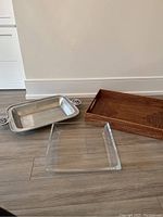 Three trays displayed on the floor: metal casserole holder with painted handles, woven rectangular tray, and shallow glass square tray.