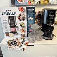 Overall view of Ninja Creami motor base, original box, three pint containers with lids, and recipe guide