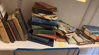 Shelf with various hardcover and softcover books stacked horizontally and vertically, showing general condition and variety of the lot.