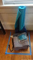 Yoga mat rolled up with blue strap standing upright next to gray yoga bolster and a small box container on the floor