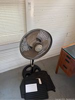 Borg floor fan with metal blades and black stand on the floor next to a wooden desk and window blinds