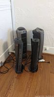Four floor fans grouped together on wooden floor by a wall, showing different models and sizes, some black and gray, with power cords visible.