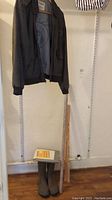 Full lot view showing jacket on rack, rain boots on floor, two wooden yardsticks leaning against wall, and storage bag on floor