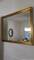 Front angled view of large gold-colored framed rectangular wall mirror hanging on white wall with a view of a round mirror reflected inside.