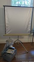 Full view of projector screen on tripod showing white projection surface, with calculator placed on base and projector cords visible