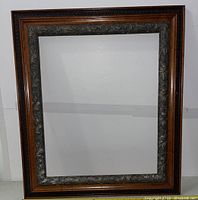 Full view of the large antique wooden picture frame showing the brown and dark wood tones with an ornate silver-toned floral inner edge.